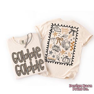 May include: Two cream-colored t-shirts. One shirt has "Gobble Gobble" in a black and white checkered pattern. The other shirt has a black border with Thanksgiving-themed graphics and the text "Gobble Til You Wobble".