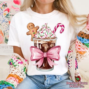 K&ouml;nnte beinhalten: Wei&szlig;es T-Shirt mit einem Grafikdruck eines Schokoladengetr&auml;nks, das mit Schlagsahne, Streuseln und einem Zuckerrohr verziert ist. Ein Lebkuchenmann lehnt sich an das Getr&auml;nk. Das Getr&auml;nk ist mit einer rosafarbenen Schleife gebunden.