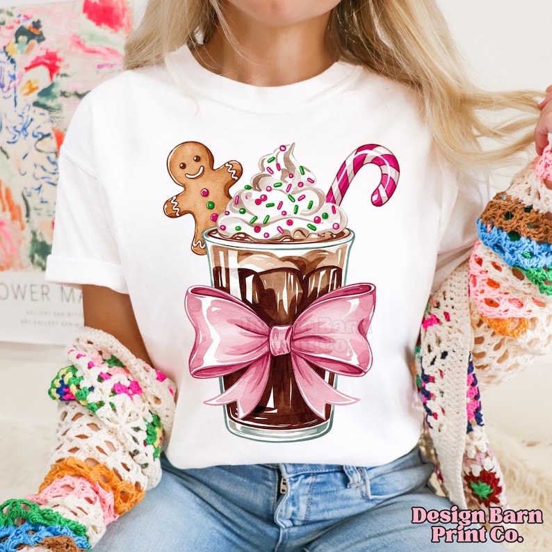 K&ouml;nnte beinhalten: Wei&szlig;es T-Shirt mit einem Grafikdruck eines Schokoladengetr&auml;nks, das mit Schlagsahne, Streuseln und einem Zuckerrohr verziert ist. Ein Lebkuchenmann lehnt sich an das Getr&auml;nk. Das Getr&auml;nk ist mit einer rosafarbenen Schleife gebunden.