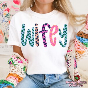 May include: White t-shirt with the word "wifey" in colorful checkered letters.