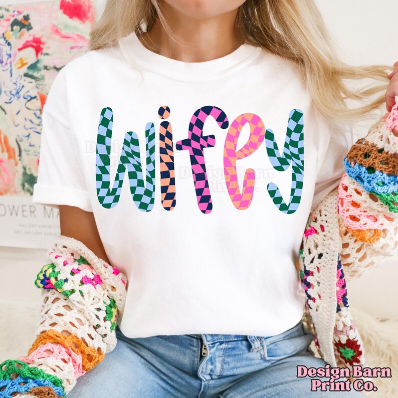 May include: White t-shirt with the word "wifey" in colorful checkered letters.