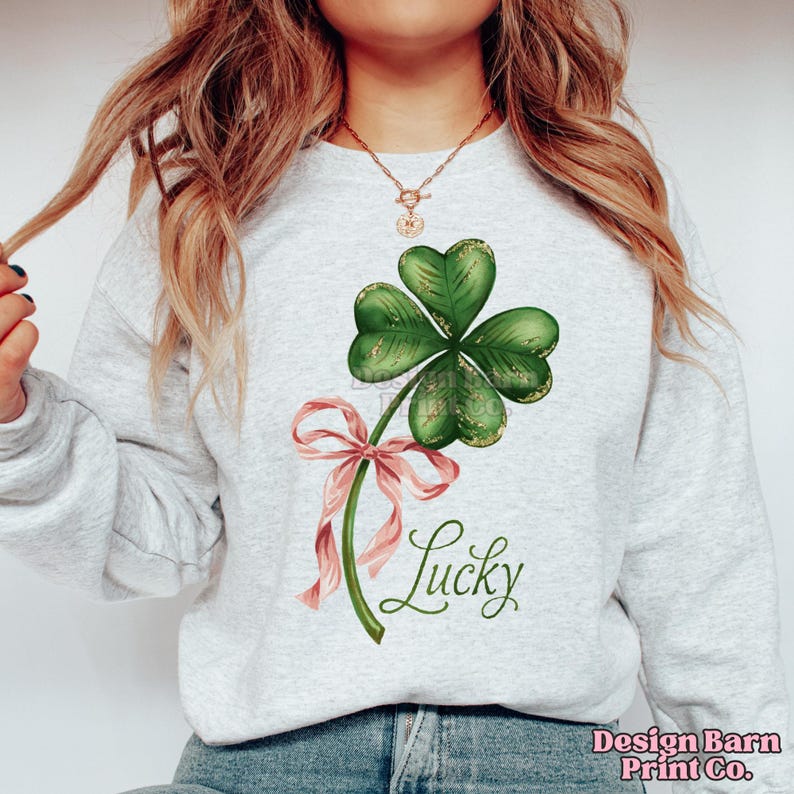 Op de afbeelding: Een grijs sweatshirt met een groene vierbladige klaver en een roze lint. De tekst "Lucky" is in cursief onder de klaver geschreven.