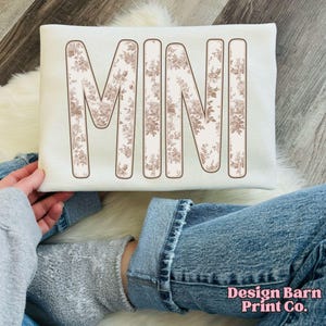 Puede incluir: Sudadera de color crema con la palabra "MINI" en letras grandes con un estampado floral. Las letras est&aacute;n delineadas en marr&oacute;n, creando un contraste con el fondo crema. La sudadera es sostenida por una persona que lleva un su&eacute;ter gris y vaqueros azules.