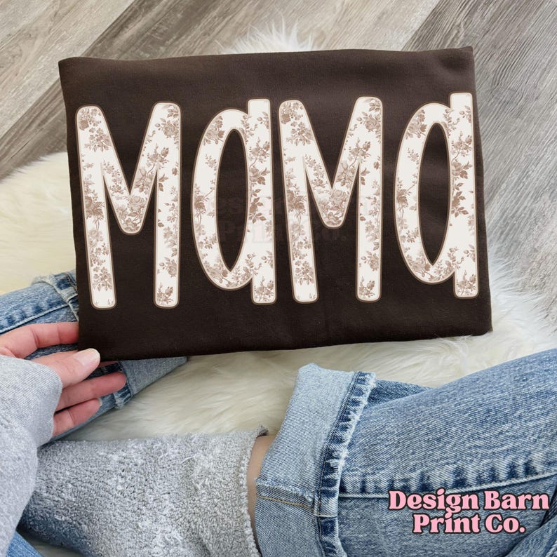 May include: A dark brown sweatshirt with the word "MAMA" in large, floral-patterned letters. The letters are outlined in a cream color, and the floral pattern features cream and brown tones. The sweatshirt is displayed on a person's lap.