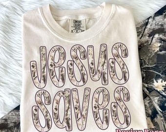 May include: Cream-colored t-shirt with the words "JESUS SAVES" in a camouflage pattern. The shirt is folded and has a tag that says "Comfort Colors". The text "Design Barn Print Co." is in the bottom right corner.