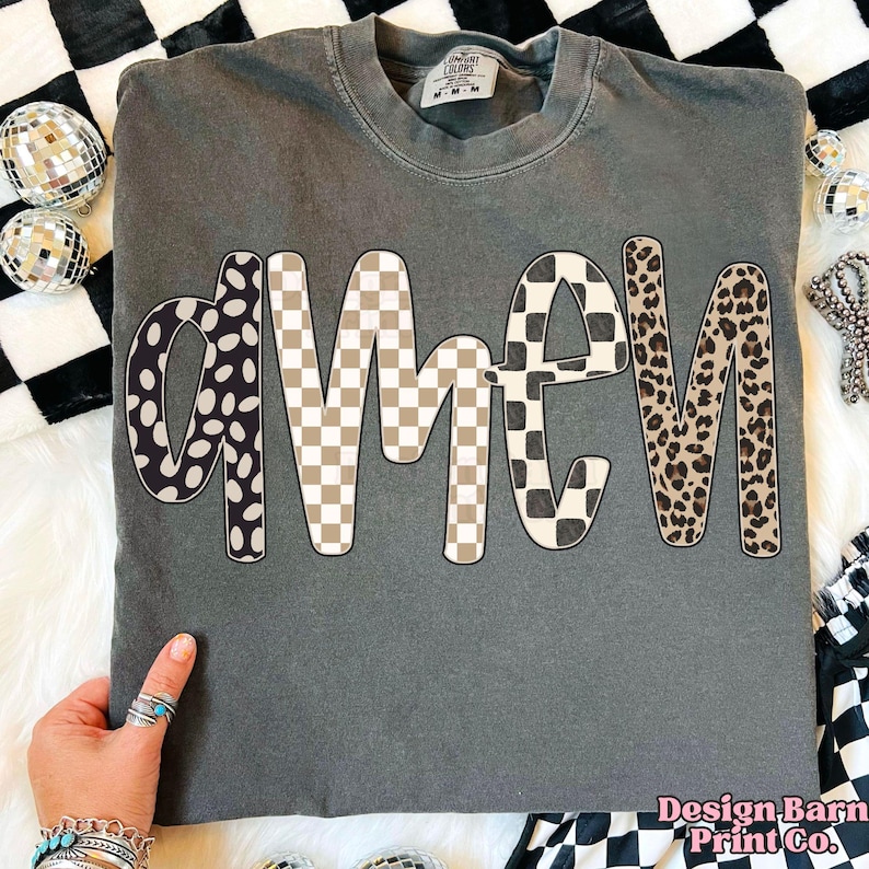 Op de afbeelding: Een grijs Comfort Colors T-shirt met het woord "AMEN" in een mix van patronen: zwart-witte stippen, bruin-wit dambord, zwart-wit dambord en luipaardprint. Het shirt wordt gepresenteerd op een wit oppervlak met discoballen en een zwart-wit dambordpatroon.