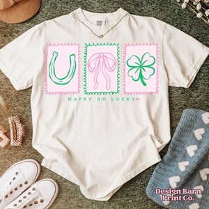 Pu&ograve; includere: T-shirt color crema con grafica rosa e verde. Il design presenta un ferro di cavallo, un fiocco e un trifoglio all'interno di cornici di francobolli rosa. Il testo "HAPPY GO LUCKY" &egrave; sotto. La maglietta &egrave; su una superficie verde.