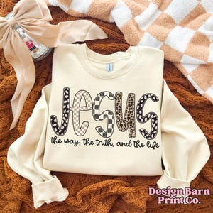 May include: A cream-coloured sweatshirt featuring the word "JESUS" in a bold, patterned font. The patterns include black and white polka dots, a checkered design, and a leopard print. The text "the way, the truth, and the life" is below the word "JESUS".