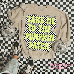 May include: A tan sweatshirt with the text "TAKE ME TO THE PUMPKIN PATCH" in a retro font. The text is outlined in black and set against a black and white checkered background. The sweatshirt is displayed on a checkered surface.