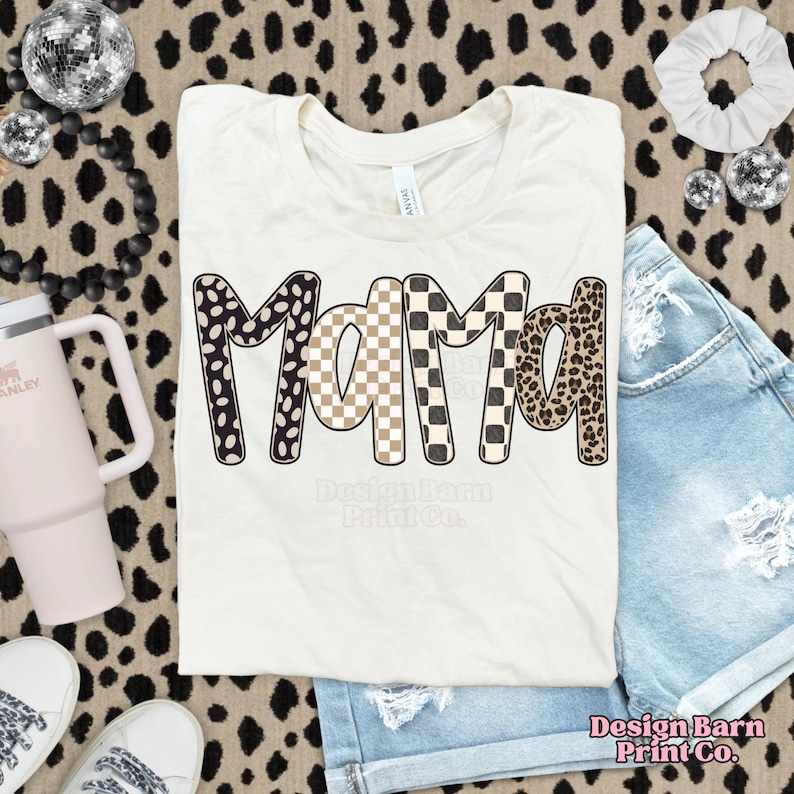 K&ouml;nnte beinhalten: Cremefarbenes T-Shirt mit dem Wort "MAMA" in einem auff&auml;lligen, gemusterten Design. Die Buchstaben weisen schwarz-wei&szlig;e Punkte, beige-wei&szlig; karierte und Leopardenmuster auf. Das Shirt wird mit Jeansshorts und Accessoires kombiniert.