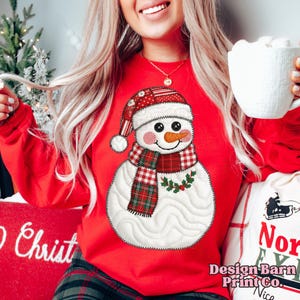 May include: A red sweatshirt featuring a snowman design. The snowman has a carrot nose, a plaid scarf, and a matching hat. The design is stitched with a quilted effect. The sweatshirt is part of a Christmas-themed collection.