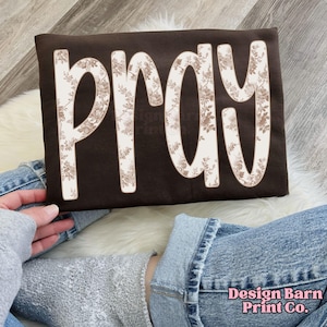 Puede incluir: Un cojín marrón con la palabra "PRAY" en letras grandes con estampado floral. Las letras son de color beige claro con un diseño floral detallado. El cojín es rectangular y parece estar hecho de una tela suave. Las palabras "Design Barn Print Co." están en la esquina inferior derecha.