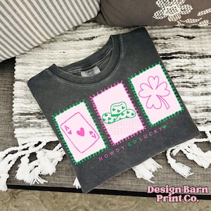 May include: Dark gray t-shirt with three pink framed graphics. The graphics include an Ace of Hearts playing card, a cowboy hat, and a four-leaf clover. The text "HOWDY GO LUCKY" is printed below the graphics.