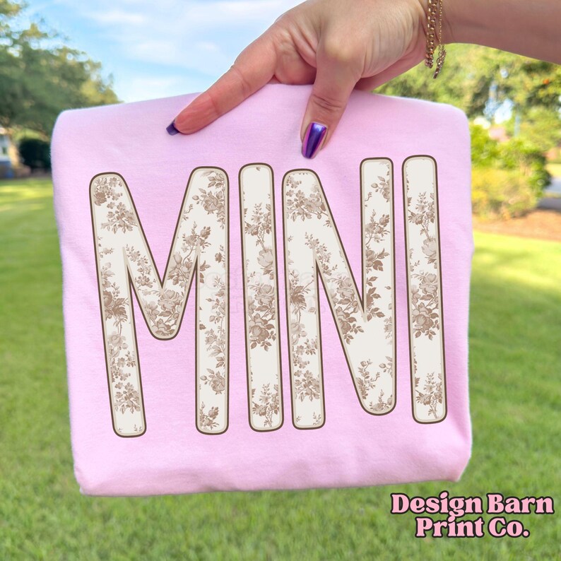 Puede incluir: Sudadera rosa claro con la palabra "MINI" en letras grandes con estampado floral en tonos beige y marr&oacute;n. La sudadera est&aacute; sujeta por una mano. El texto "Design Barn Print Co." es visible en la esquina inferior derecha.