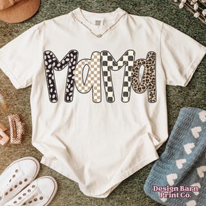 May include: Cream-colored t-shirt with the word "Mama" in a bold, patterned design. The letters feature black and white polka dots, a tan and white checkerboard, and a leopard print. The shirt is styled with white sneakers and a necklace.