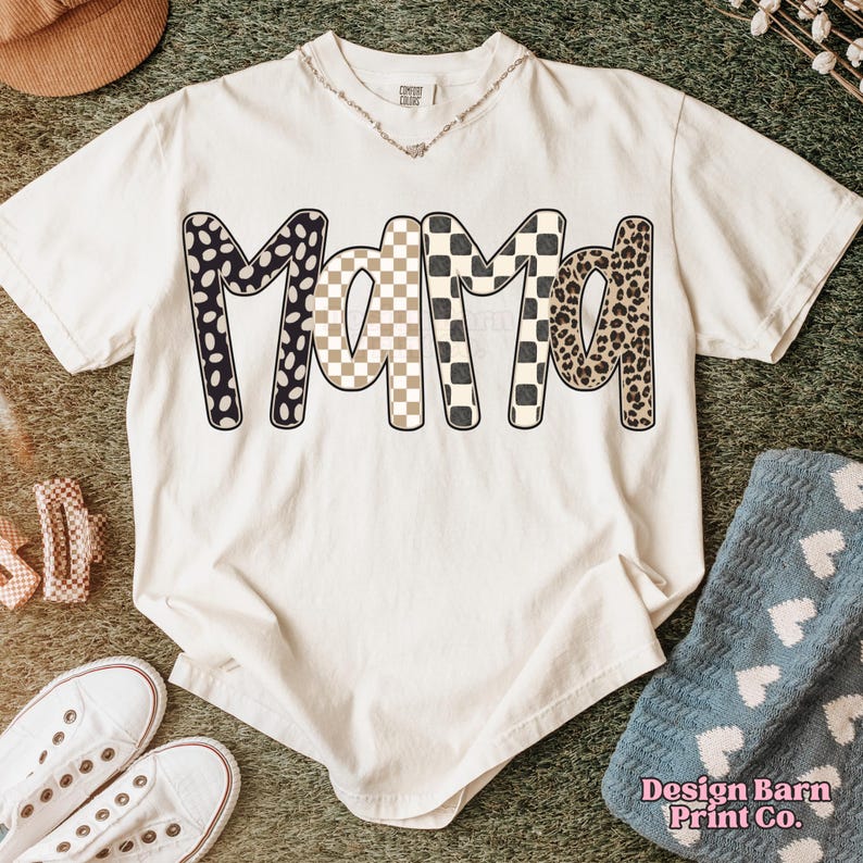 K&ouml;nnte beinhalten: Cremefarbenes T-Shirt mit dem Wort "Mama" in einem auff&auml;lligen, gemusterten Design. Die Buchstaben zeigen schwarze und wei&szlig;e Tupfen, ein beige-wei&szlig;es Schachbrettmuster und einen Leopardenprint. Das Shirt wird mit wei&szlig;en Turnschuhen und einer Halskette gestylt.