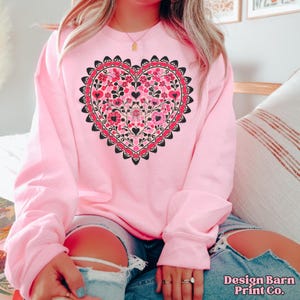 K&ouml;nnte beinhalten: Ein hellrosa Sweatshirt mit einem gro&szlig;en Herzdesign. Das Herz ist mit rosa und roten Blumenmustern gef&uuml;llt, die schwarz umrandet sind. Das Sweatshirt hat lange &Auml;rmel und einen Rundhalsausschnitt. Der Text "Design Barn Print Co." ist unten rechts sichtbar.