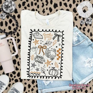 May include: Cream-colored t-shirt with a Thanksgiving-themed graphic. The design includes a pie slice, turkey, pumpkin, and the text "Thanksgiving" and "Gobble Til You Wobble" in a black, brown, and gold color scheme. The shirt is paired with denim shorts.