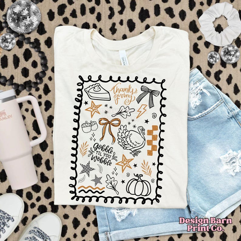 May include: Cream-colored t-shirt with a Thanksgiving-themed graphic. The design includes a pie slice, turkey, pumpkin, and the text "Thanksgiving" and "Gobble Til You Wobble" in a black, brown, and gold color scheme. The shirt is paired with denim shorts.