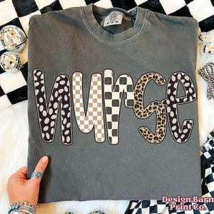 May include: A gray Comfort Colors t-shirt with the word "NURSE" in a mix of patterns: black and white polka dots, tan and white checkerboard, and leopard print. The shirt is displayed on a white surface with black and white checkered fabric.