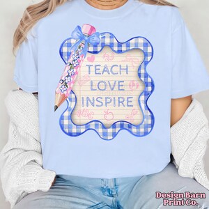 Pode incluir: Camiseta azul clara com um design tem&aacute;tico para professores. O gr&aacute;fico inclui um l&aacute;pis com detalhes florais, uma moldura xadrez azul e as palavras "Teach Love Inspire". Elementos adicionais incluem cora&ccedil;&otilde;es, ma&ccedil;&atilde;s e tesouras.