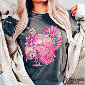 May include: A dark gray t-shirt with a pink and iridescent glitter turkey design. The turkey is wearing a bow and has a disco ball body. The shirt has the text "Design Barn Print Co." on the bottom.