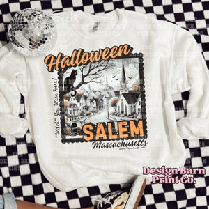K&ouml;nnte beinhalten: Wei&szlig;es Sweatshirt mit einer Grafik im Vintage-Stil, die eine schwarze Katze zeigt, die auf einem Baumzweig sitzt, vor einer gruseligen Stadt mit einer Kirche. Der Text "Halloween" befindet sich oben auf der Grafik und "Salem, Massachusetts" unten. Der Text "Witch you were here!" befindet sich auf der linken Seite der Grafik.