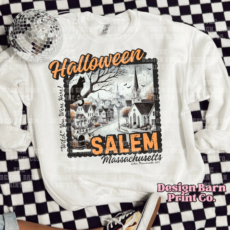 K&ouml;nnte beinhalten: Wei&szlig;es Sweatshirt mit einer Grafik im Vintage-Stil, die eine schwarze Katze zeigt, die auf einem Baumzweig sitzt, vor einer gruseligen Stadt mit einer Kirche. Der Text "Halloween" befindet sich oben auf der Grafik und "Salem, Massachusetts" unten. Der Text "Witch you were here!" befindet sich auf der linken Seite der Grafik.