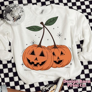 K&ouml;nnte beinhalten: Wei&szlig;er Pullover mit einem Aufdruck von zwei orangefarbenen K&uuml;rbissen in Form von Kirschen mit gr&uuml;nen Stielen und Bl&auml;ttern. Die K&uuml;rbisse haben Jack-o'-Lantern-Gesichter. Der Hintergrund ist wei&szlig; mit kleinen wei&szlig;en Sternen.