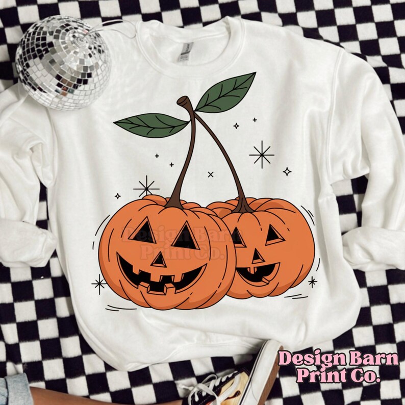 K&ouml;nnte beinhalten: Wei&szlig;er Pullover mit einem Aufdruck von zwei orangefarbenen K&uuml;rbissen in Form von Kirschen mit gr&uuml;nen Stielen und Bl&auml;ttern. Die K&uuml;rbisse haben Jack-o'-Lantern-Gesichter. Der Hintergrund ist wei&szlig; mit kleinen wei&szlig;en Sternen.