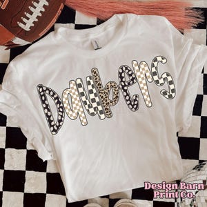 May include: A white t-shirt with the word "Daybem's" in a mix of patterns, including polka dots, leopard print, and checkered designs. The shirt is on a black and white checkered surface. The text "Design Barn Print Co." is visible.