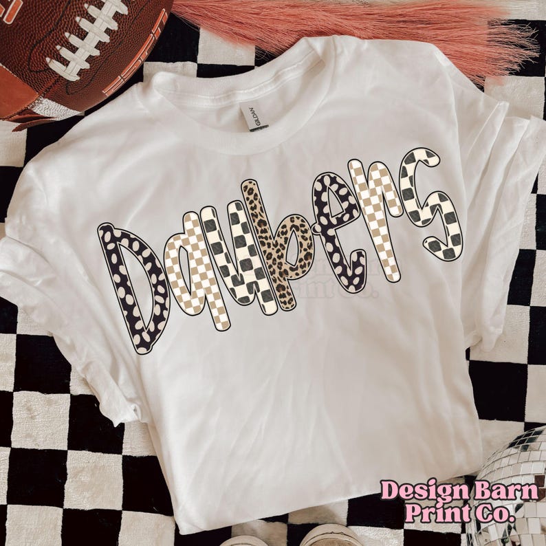 May include: A white t-shirt with the word "Daybem's" in a mix of patterns, including polka dots, leopard print, and checkered designs. The shirt is on a black and white checkered surface. The text "Design Barn Print Co." is visible.