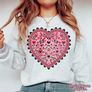 K&ouml;nnte beinhalten: Hellgraues Sweatshirt mit einem gro&szlig;en Herzdesign, gef&uuml;llt mit rosa Blumen, schwarzen Herzen und einem roten Rand. Das Herz ist mittig auf der Brust platziert. Der Text "Design Barn Print Co." befindet sich unten rechts.