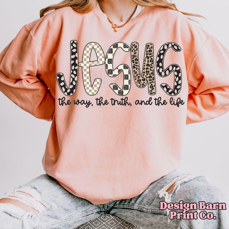 May include: A light pink sweatshirt with the word "JESUS" in a bold, patterned font. The letters feature a mix of black and white polka dots, checkered patterns, and leopard print. Below the word is the phrase "the way, the truth, and the life."