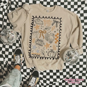 Puede incluir: Sudadera beige con un dise&ntilde;o de Acci&oacute;n de Gracias en negro y naranja. El dise&ntilde;o incluye un pavo, una calabaza, un pastel y el texto "Gobble Til You Wobble" y "Thanksgiving". La sudadera est&aacute; sobre una superficie a cuadros blancos y negros.
