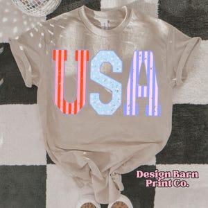 May include: A beige t-shirt with the word "USA" printed on the front. The letters are in a vintage style with red, white, and blue stripes and stars. The text "Design Barn Print Co." is printed in pink at the bottom of the shirt.