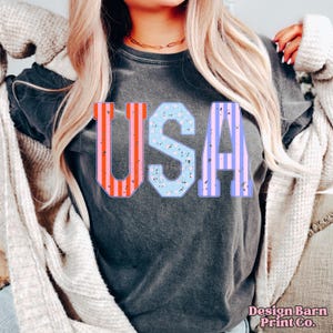 May include: A dark gray t-shirt with the word "USA" printed on the front. The letters are in a vintage style with red, blue, and pink stripes and stars.
