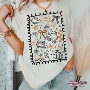 May include: Cream-colored t-shirt with a Thanksgiving-themed graphic. The design includes a pie slice, bow, turkey, stars, and the text "Thanksgiving" and "Gobble Til You Wobble". The graphic is outlined in black.