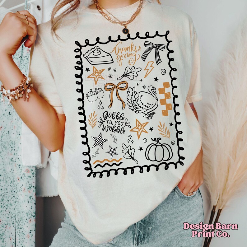 May include: Cream-colored t-shirt with a Thanksgiving-themed graphic. The design includes a pie slice, bow, turkey, stars, and the text "Thanksgiving" and "Gobble Til You Wobble". The graphic is outlined in black.