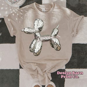 May include: A light brown t-shirt with a silver disco ball balloon animal design. The design is of a dog with a long tail. The shirt has a crew neck and short sleeves. The text "Design Barn Print Co." is printed on the bottom right corner of the shirt.