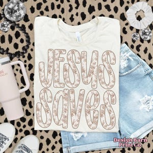 May include: A cream-coloured t-shirt with the words "JESUS SAVES" in large, floral-patterned letters. The shirt is paired with denim shorts, white trainers, and a pink tumbler. A white scrunchie and disco balls are also in the image.