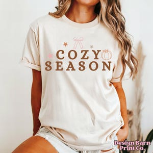 May include: Cream-colored t-shirt with the words "COZY SEASON" in brown letters. The design includes a pink bow, stars, and a pumpkin graphic. The shirt is a short-sleeved, casual style.