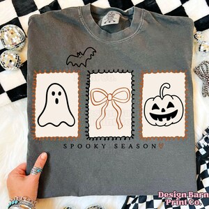 May include: A gray long-sleeve shirt with a Halloween theme. The shirt features a bat, a ghost, a bow, and a jack-o'-lantern design. The words "Spooky Season" are printed below the designs. The shirt is from Design Barn Print Co.