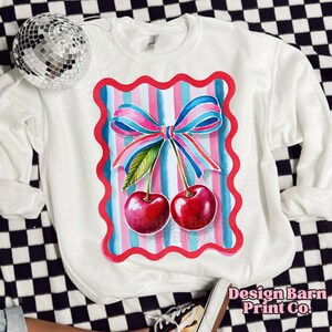 May include: White sweatshirt with a red, pink, and blue striped design featuring two cherries with green stems and leaves. The cherries are surrounded by a pink bow.