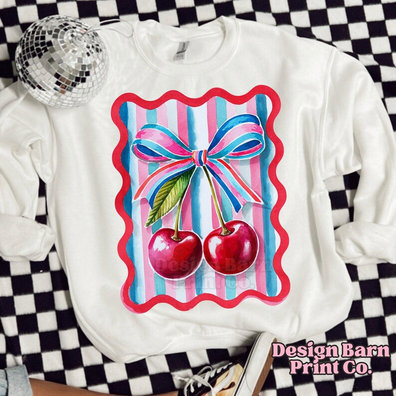May include: White sweatshirt with a red, pink, and blue striped design featuring two cherries with green stems and leaves. The cherries are surrounded by a pink bow.