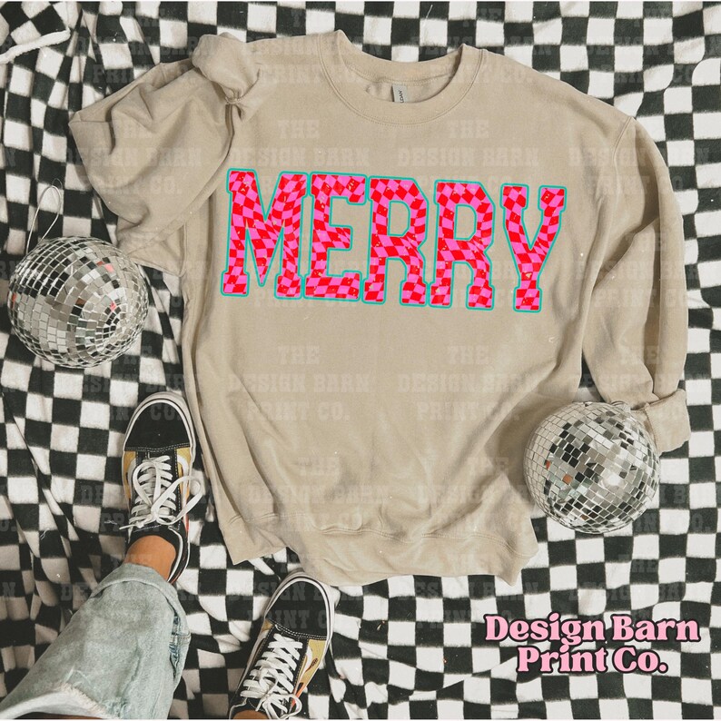 May include: A beige sweatshirt with a pink and blue "MERRY" graphic design. The graphic is made up of pink and red checkered squares. The sweatshirt is laying on a black and white checkered surface.