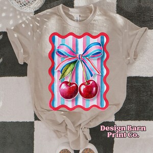 May include: A beige t-shirt with a red, white, and blue striped design featuring a pink bow and two red cherries. The design is framed in a red scalloped border.