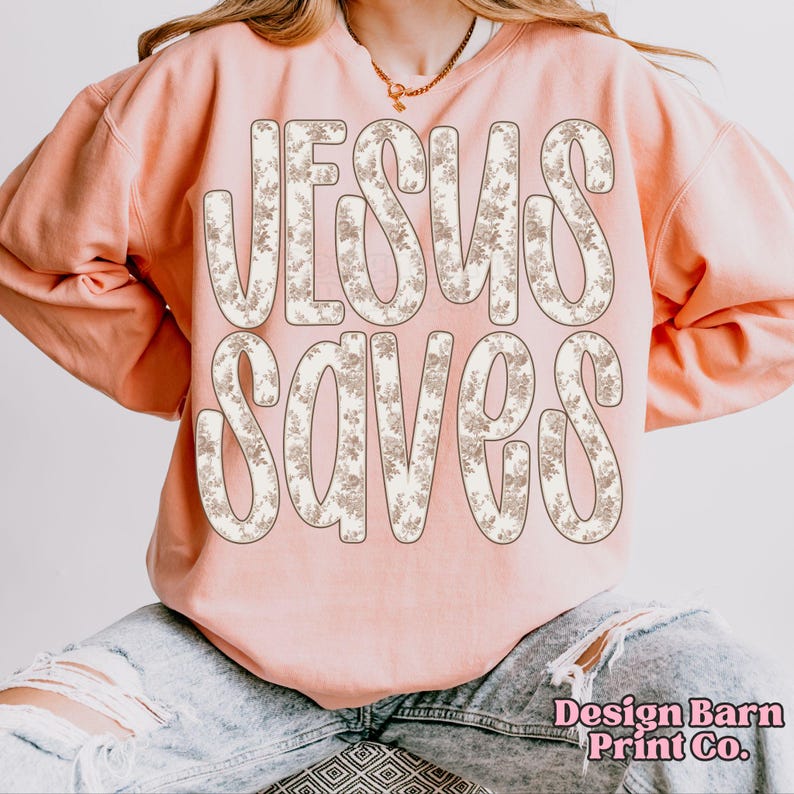 May include: A pale pink sweatshirt featuring the text "JESUS SAVES" in large letters filled with a floral pattern. The sweatshirt has a crew neck and long sleeves. The words "Design Barn Print Co." are printed in the bottom right corner.