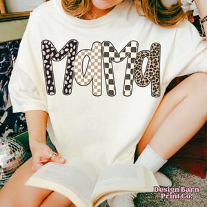 K&ouml;nnte beinhalten: Cremefarbenes T-Shirt mit dem Wort "Mama" in einem auff&auml;lligen, gemusterten Design. Die Buchstaben zeigen eine Mischung aus schwarzen und wei&szlig;en Punkten, karierten Mustern in Beige und Wei&szlig;, karierten Mustern in Schwarz und Wei&szlig; und Leopardenmuster. Das Shirt ist von Design Barn Print Co.