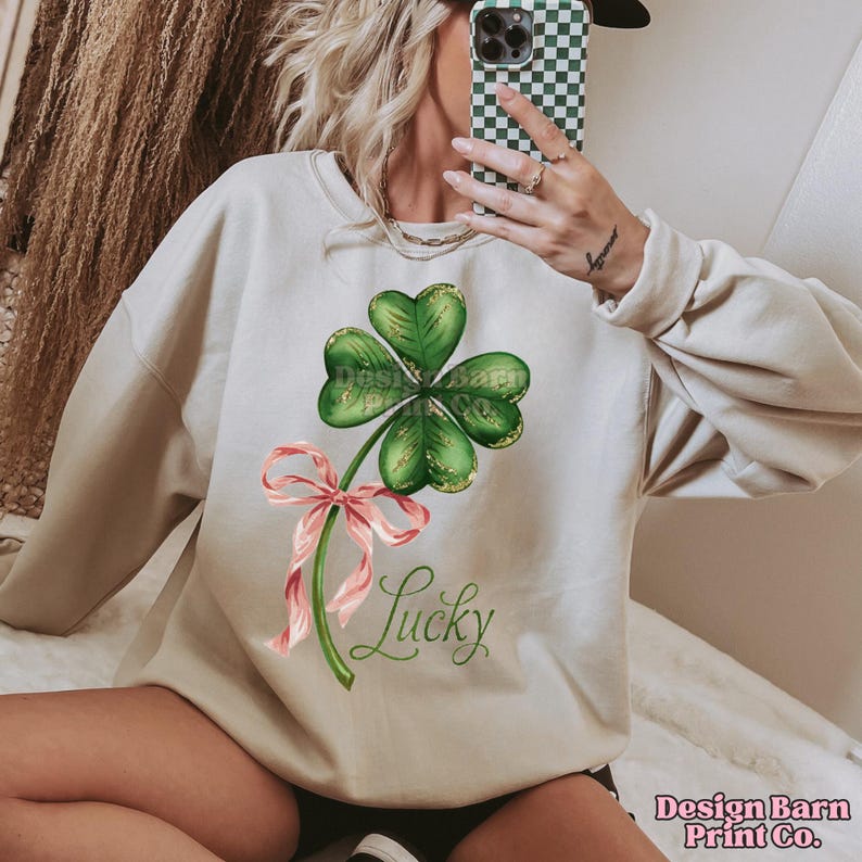 Op de afbeelding: Een licht beige sweatshirt met een groene vierbladige klaver met gouden glitteraccenten en een roze lint. De tekst "Lucky" is onder de klaver gedrukt.